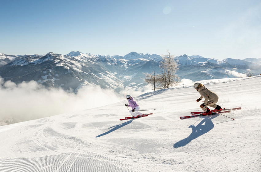 Zwei Skifahrerinnen fahren bei Sonne die Piste hinab mit Bergpanorama im Hintergrund | © Großarler Bergbahnen GmbH & Co KG
