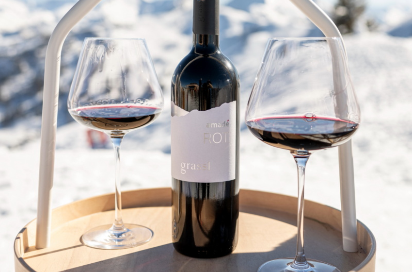 Weinflasche steht zwischen zwei gefüllten Weingläsern auf kleinem Tisch im Schnee vor alpiner Berglandschaft | © Großarler Bergbahnen GmbH & Co KG