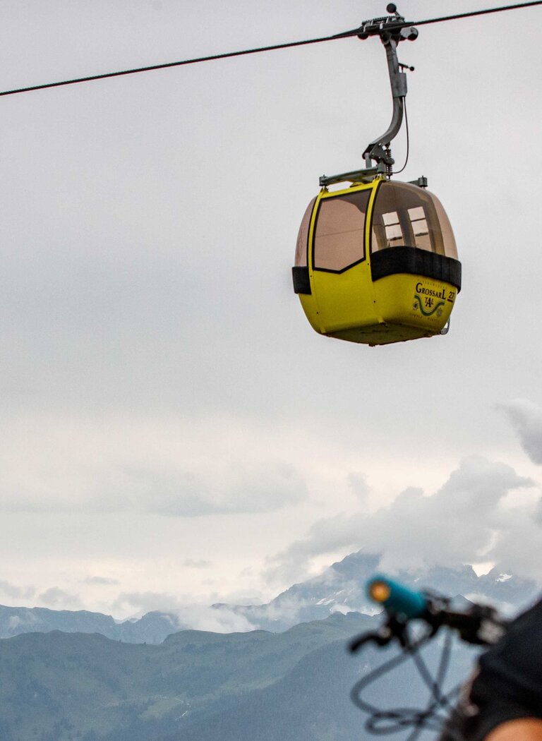 Radfahrer mit Helm blickt auf gelbe Gondel der Großarlbahn vor bewölktem Himmel und alpinem Bergpanorama | © Grossarler Bergbahnen GmbH & Co KG