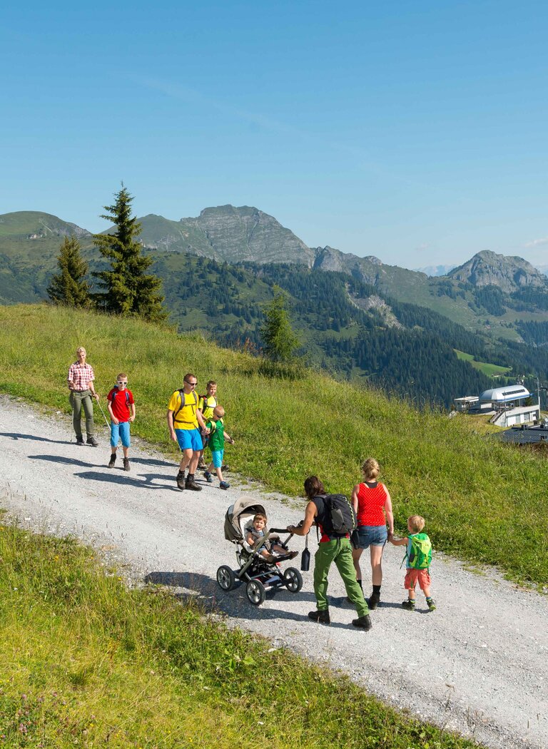 Familien mit Kindern und Kinderwagen auf sonnigem Almweg in Großarl, daneben gelbe Gondeln und Weitblick ins Tal | © Grossarler Bergbahnen GmbH & Co KG
