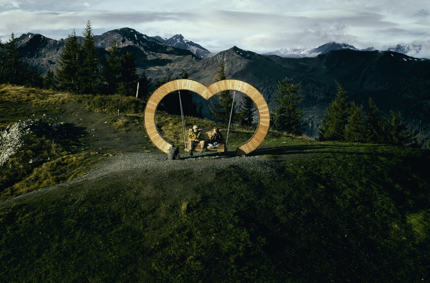 Two people on a heart-shaped swing bench with mountain views, forest, and autumn landscape in the background. | © Grossarler Bergbahnen GmbH & Co KG