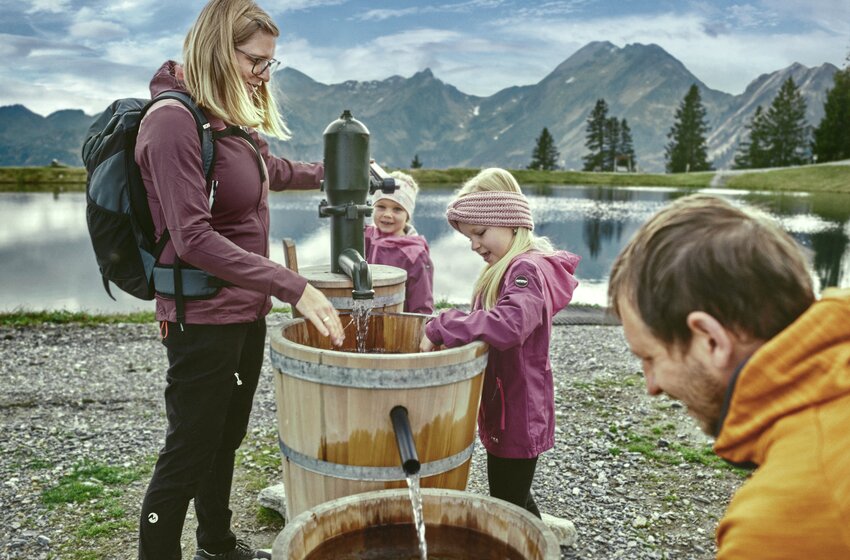 Family at a water pump with wooden barrels by a mountain lake, kids in jackets playing, smiling parents. | © Grossarler Bergbahnen GmbH & Co KG
