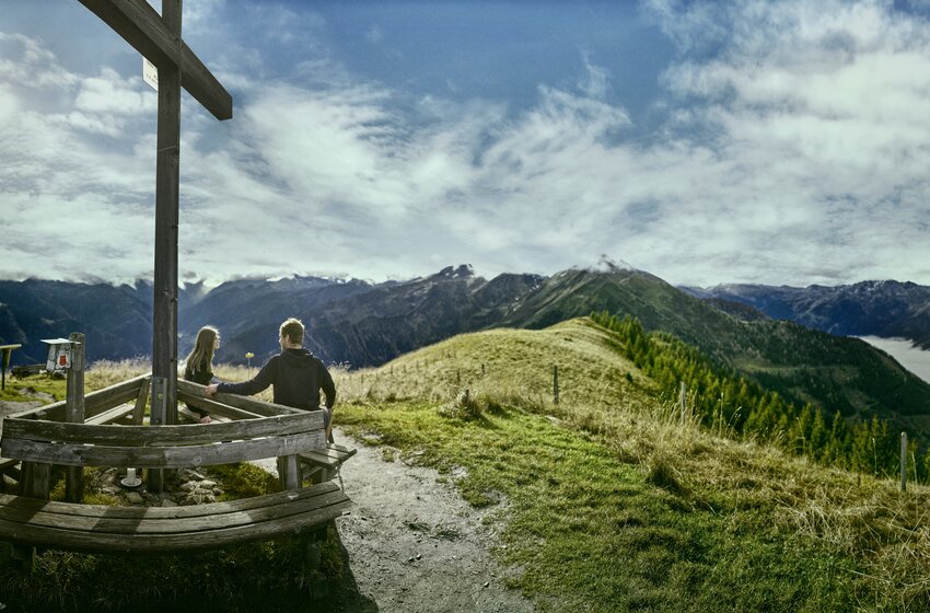 Couple at summit cross enjoying distant alpine valley views | © Grossarler Bergbahnen GmbH & Co KG Couple sitting at summit cross with benches, overlooking mountain landscape and fog-covered valleys. | © Grossarler Bergbahnen GmbH & Co KG