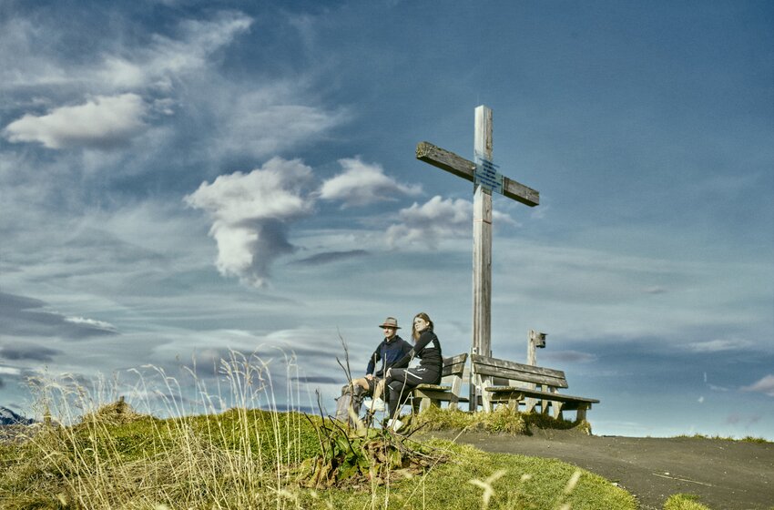 Two hikers sit on a bench beside a summit cross with grassy hillside, trail and dramatic cloud-filled sky. | © Grossarler Bergbahnen GmbH & Co KG