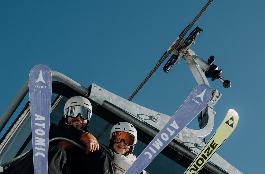 Zwei Skifahrer sitzen mit angeschnallten Skiern im Sessellift unter klarem Himmel und blicken nach unten. | © Elisabeth Toferer