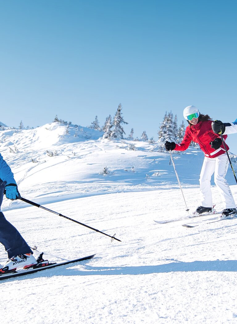 Drei Skifahrer fahren nebeneinander auf einer sonnigen, verschneiten Piste mit Sesselliften im Hintergrund in Ski amadé. | © Ski amadé GmbH