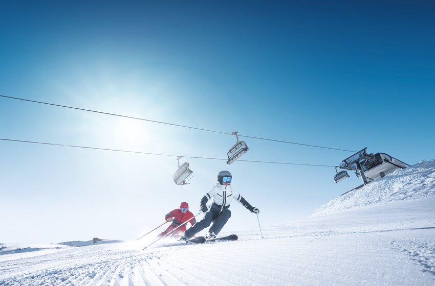 Two skiers carving dynamically under a sunny chairlift ride | © Ski amadé GmbH Two skiers carve on a freshly groomed slope beneath a chairlift under a bright blue sky and glowing morning sun. | © Ski amadé GmbH