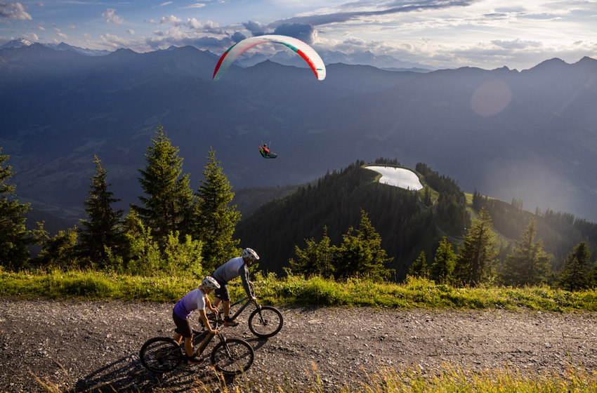 Two mountain bikers ride on a gravel trail on a slope, above a paraglider flies, and a mountain lake lies in the valley | © Gasteiner Bergbahnen AG